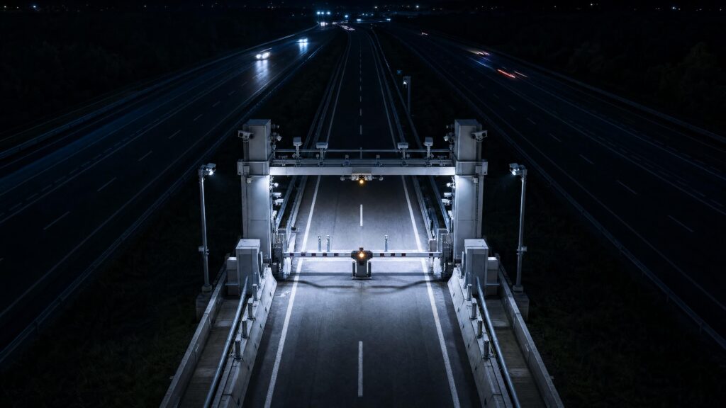 An empty motorway toll plaza at night, its lanes lit white and running away toward distant traffic
