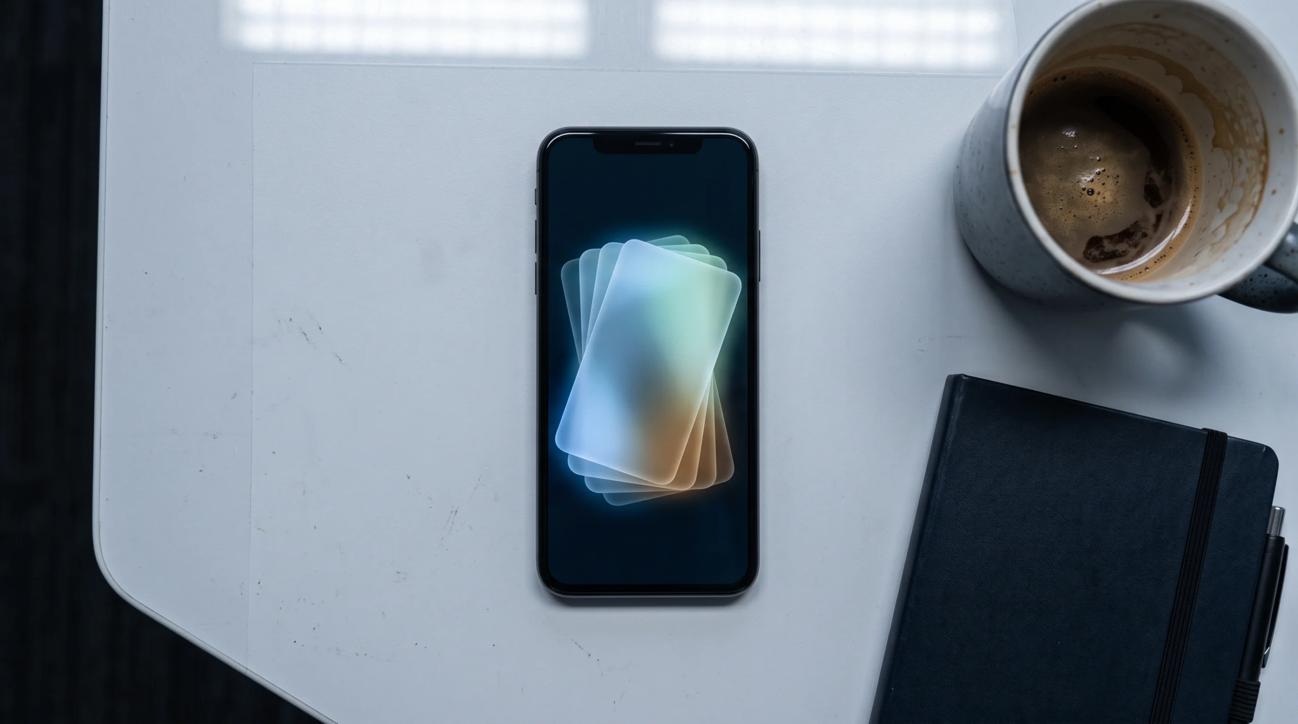 A close-up still life on a plain desk: a smartphone lies face-up showing a softly glowing stack of overlapping translucent cards fanned across the screen, each one…